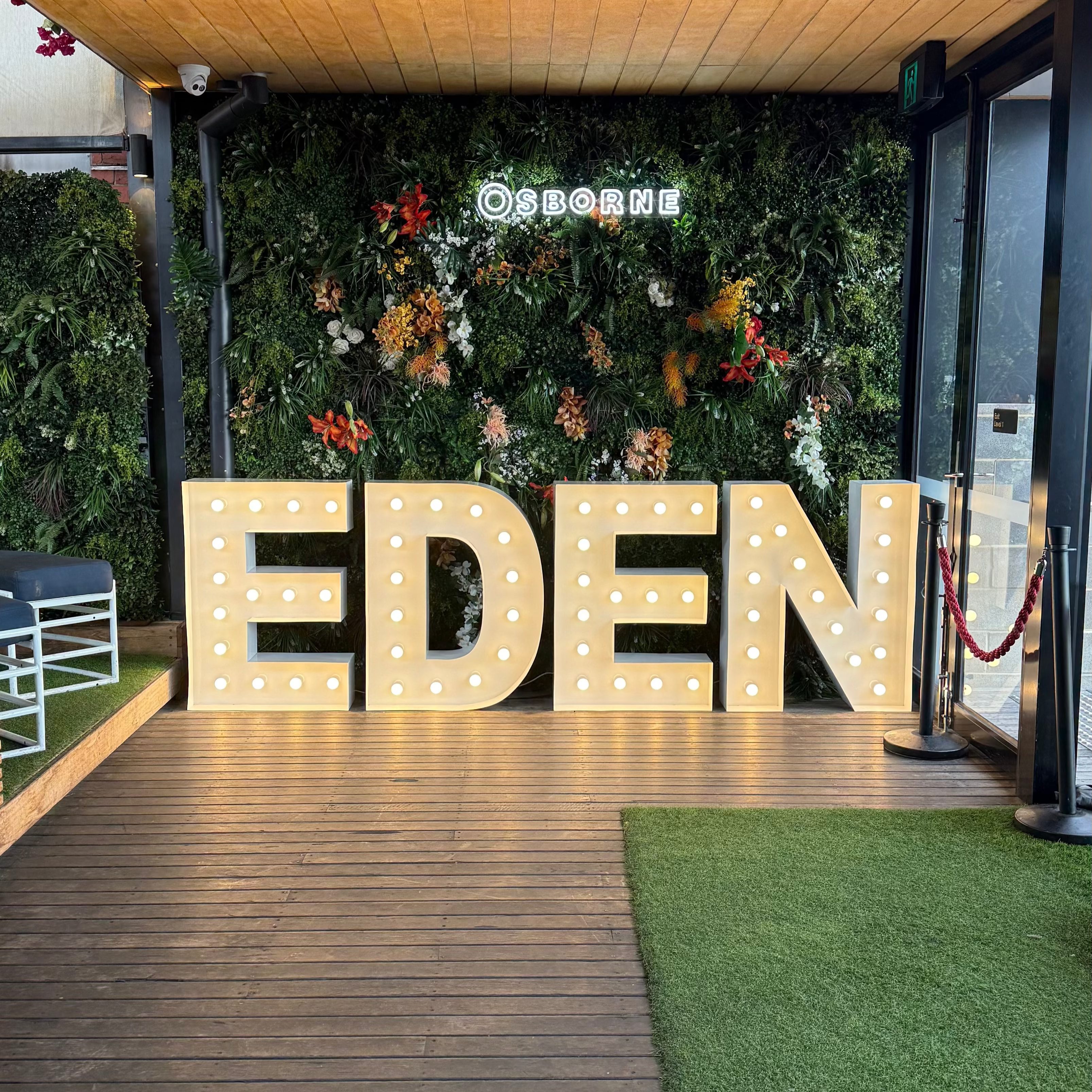 Large illuminated letters spelling 'EDEN' on a wooden deck with greenery in the background.
