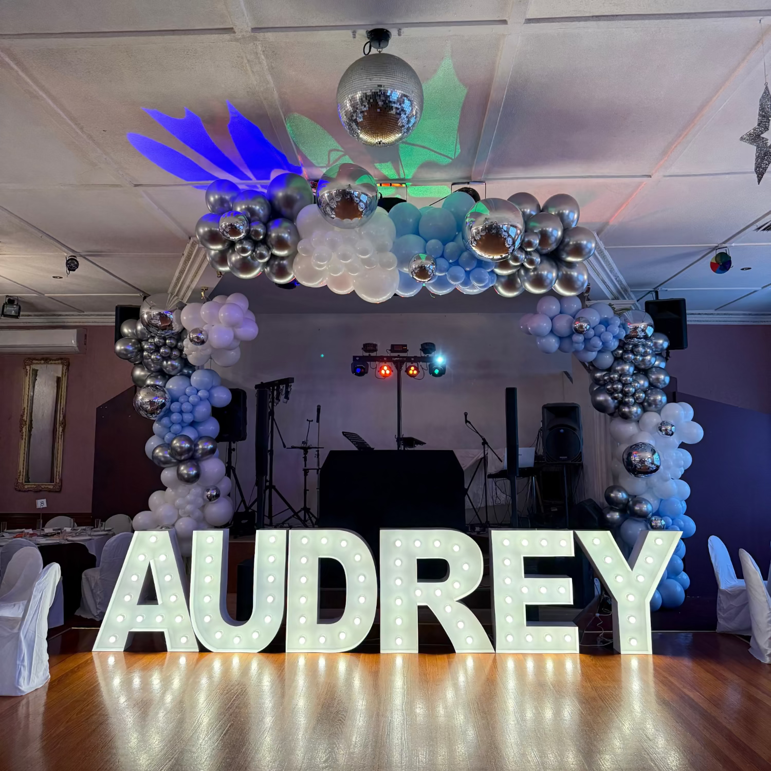 Decorative arch with balloons and 'AUDREY' letters in a room with disco lights.