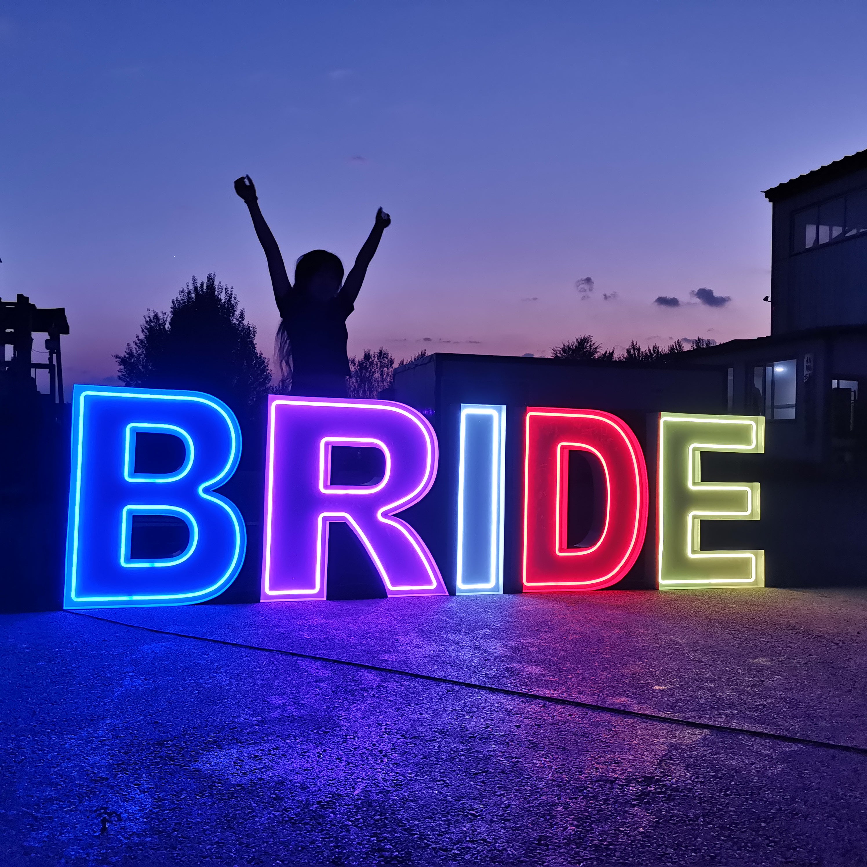 Person with arms raised behind a colorful 'BRIDE' neon letters at dusk.
