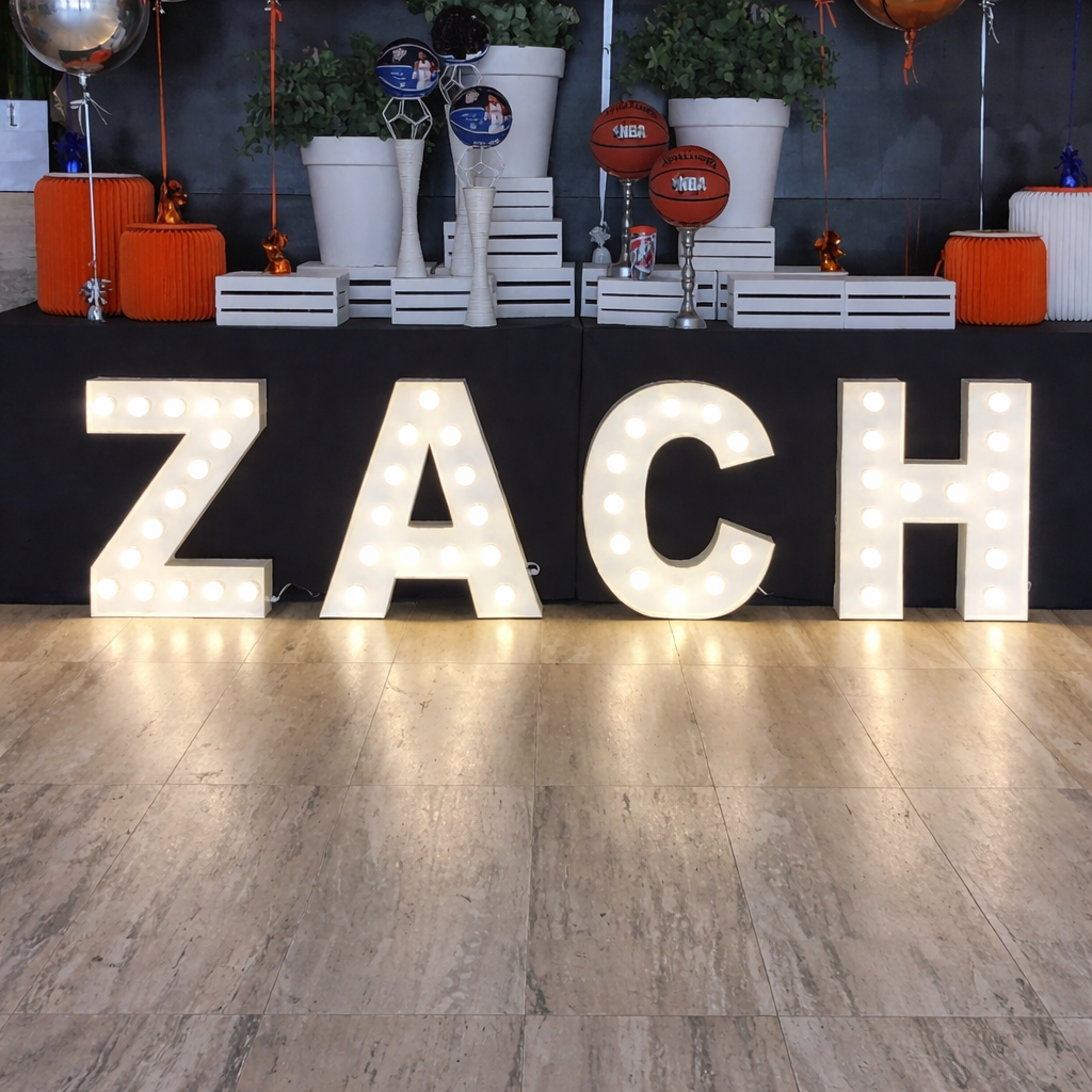 Large illuminated letters spelling 'ZACH' on a wooden floor with decorative items in the background.