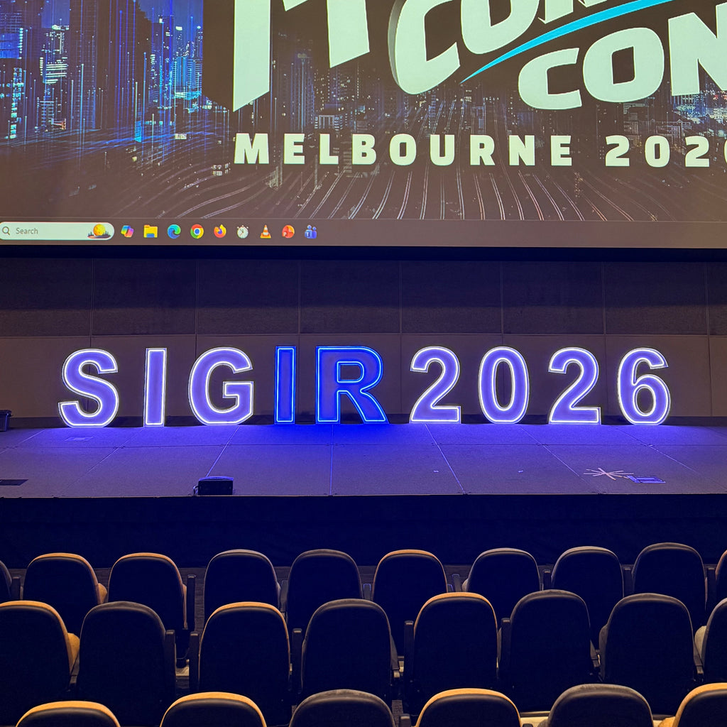 A conference stage with large blue LED numbers reading 'SIGIR 2026' and a screen behind showing 'MELBOURNE 2026' and partial event branding. Rows of dark audience seats are visible in the foreground.