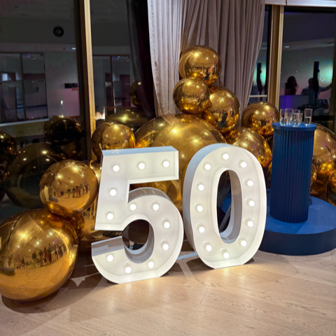 Decorative setup with gold balloons and a large illuminated '50' sign indoors.