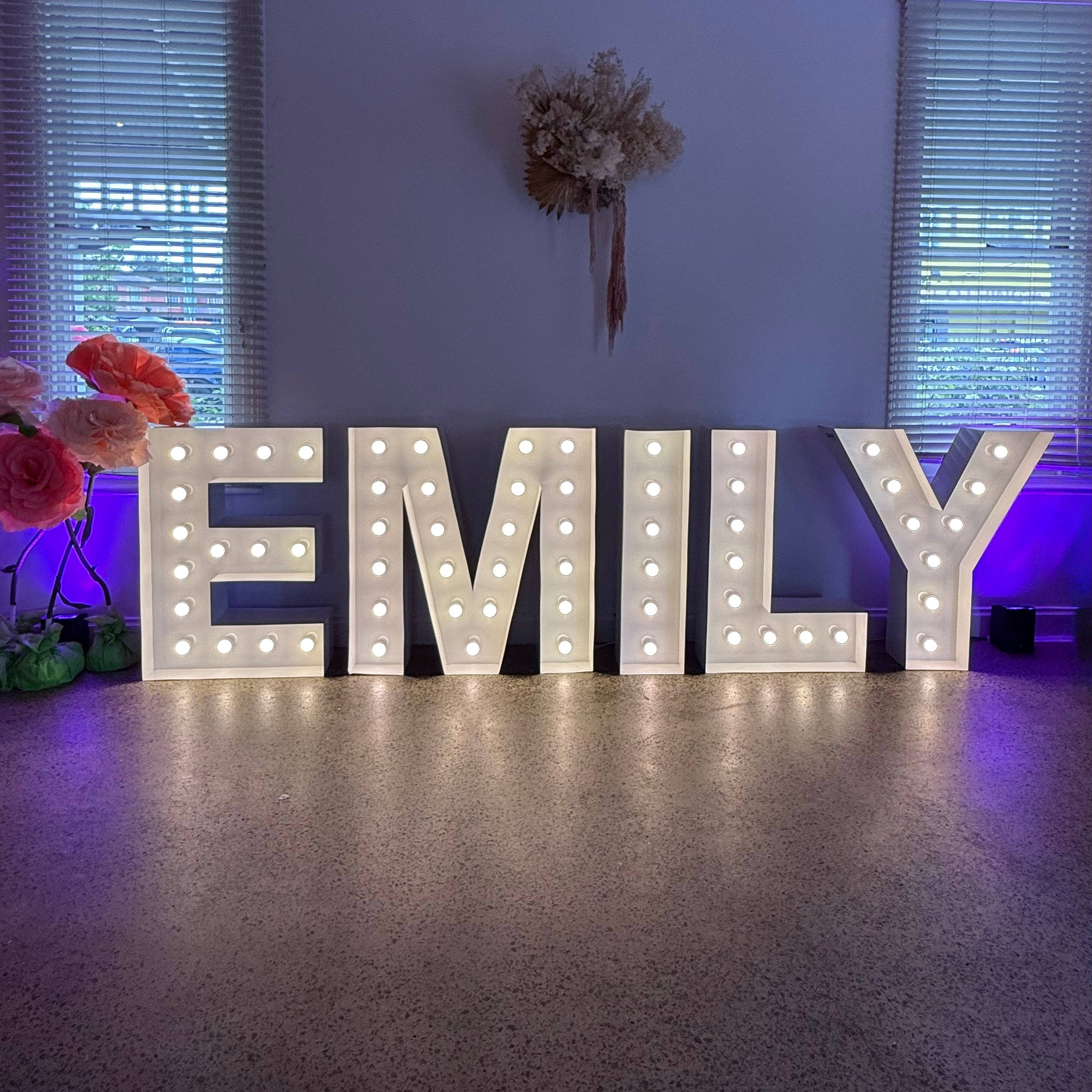 Decorative letters spelling 'EMILY' with lights on a floor in a room with windows and a floral arrangement.